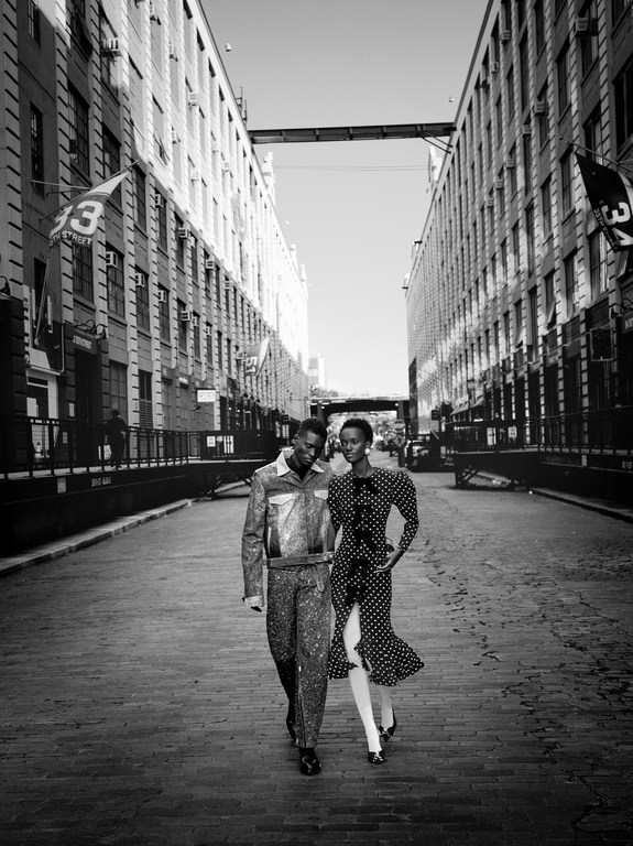 A man and a woman stroll down a street lined with brick buildings in a black and white fashion photograph with urban style.
