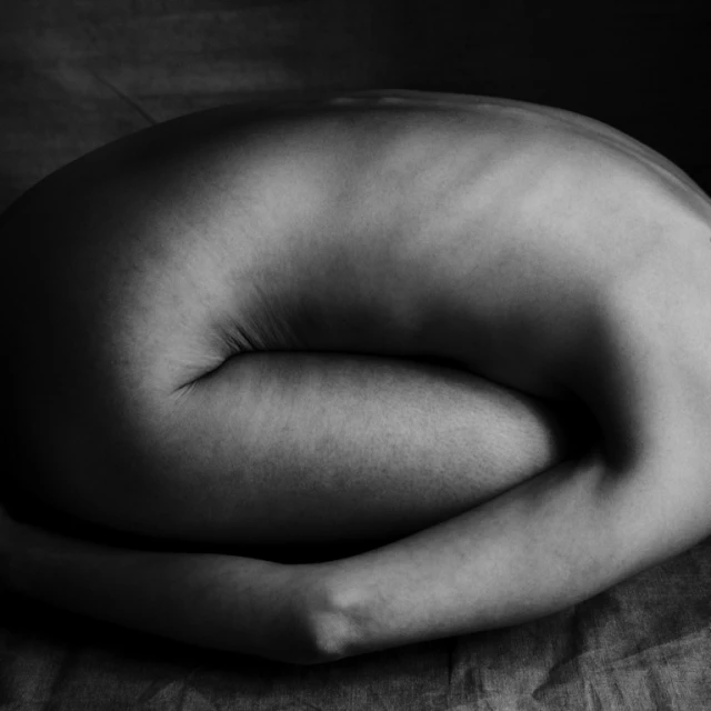 Monochrome fine art nude photography of a woman curled up, emphasizing body form and shadows against a textured background in a studio setting.