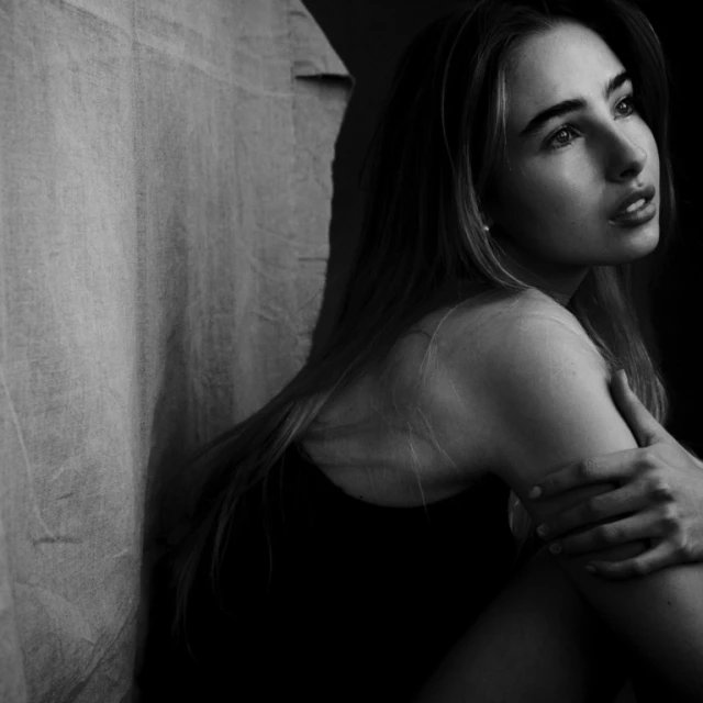 Black and white portrait of a young adult woman with long hair, resting her arms, in a dimly lit setting near a draped fabric.