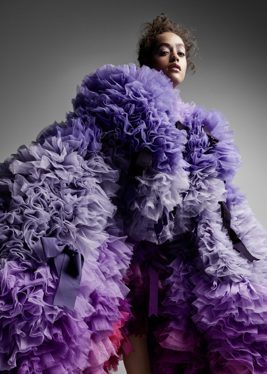 Woman wearing a ruffled purple gown poses in front of a gray backdrop. Fashion studio shot of model with neutral expression.