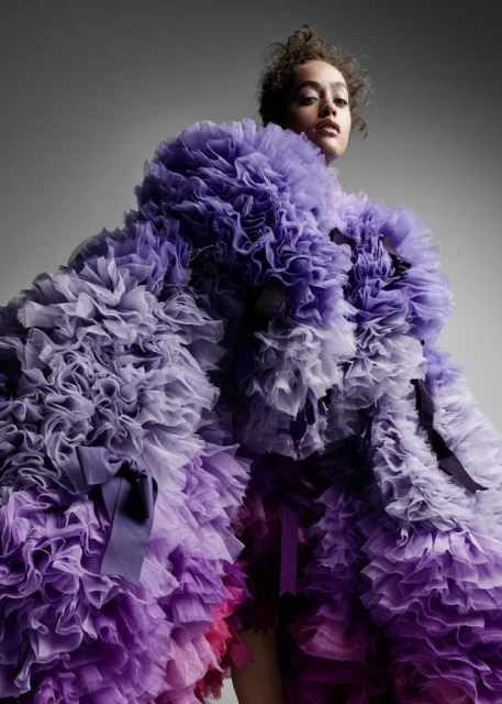 Woman wearing a ruffled purple gown poses in front of a gray backdrop. Fashion studio shot of model with neutral expression.