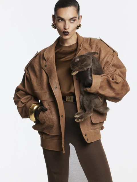 Brunette adult woman in tan jacket poses indoors holding brown rabbit, wearing gold cuff bracelet and earrings against a white backdrop.
