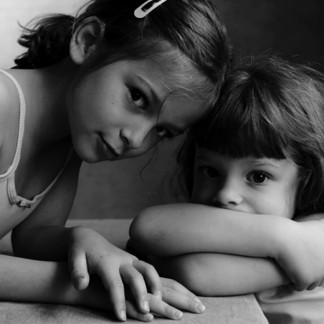Black and white portrait of two young girls indoors, one with a hair clip, in a close, loving pose, looking at the camera with soft lighting.