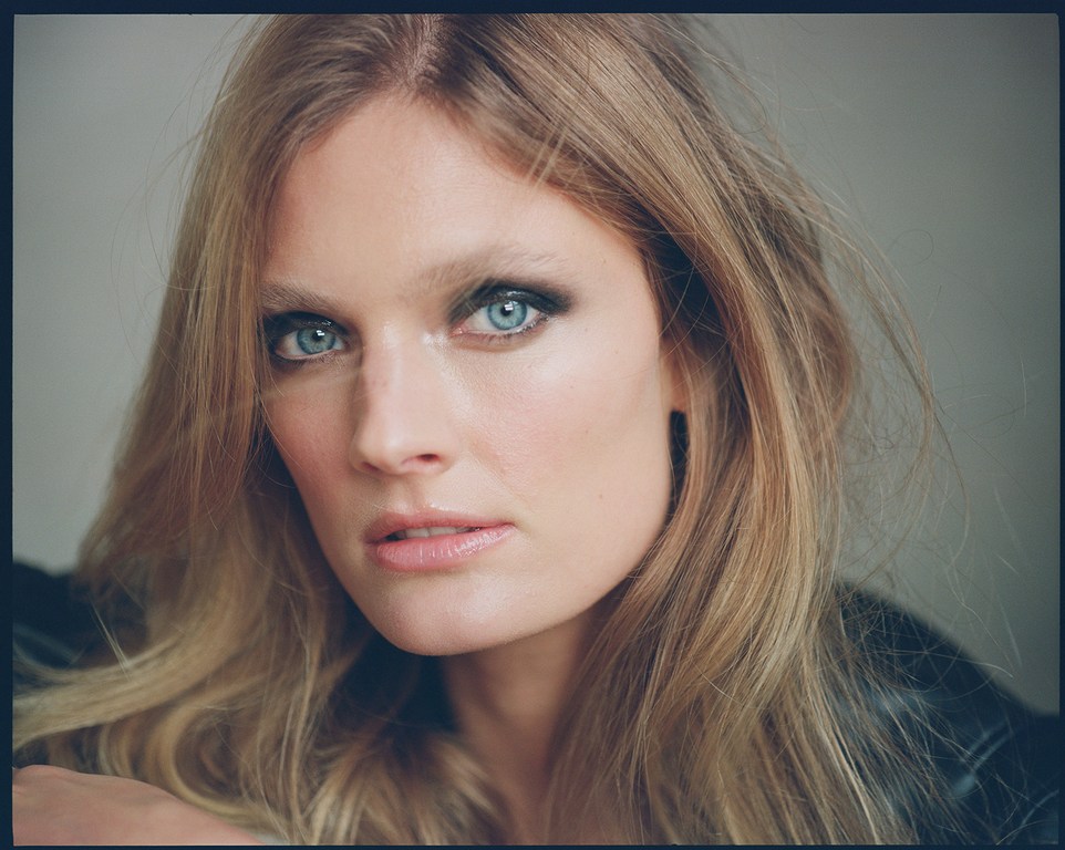 Close-up of a blonde adult woman with light skin, blue eyes, and prominent makeup, wearing a black leather jacket against a neutral background.
