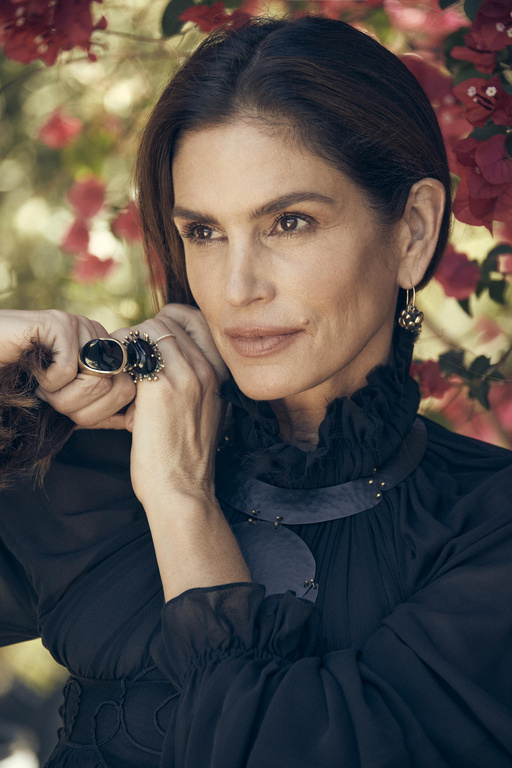 Close-up portrait of a mature woman with dark hair, wearing a black dress, statement ring and earrings, posing in front of red flowers.