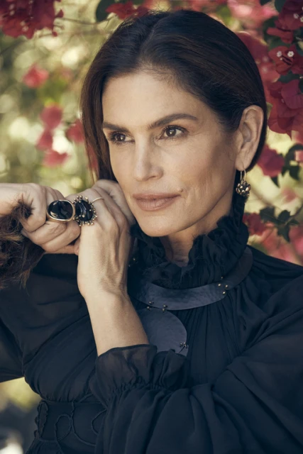 Close-up portrait of a mature woman with dark hair, wearing a black dress, statement ring and earrings, posing in front of red flowers.