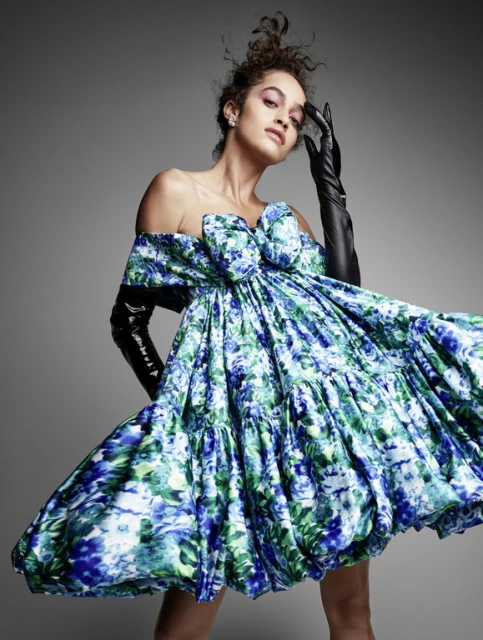 Elegant woman in flowing floral mini dress with long black gloves posing in a studio with a smooth grey backdrop for a fashion editorial.