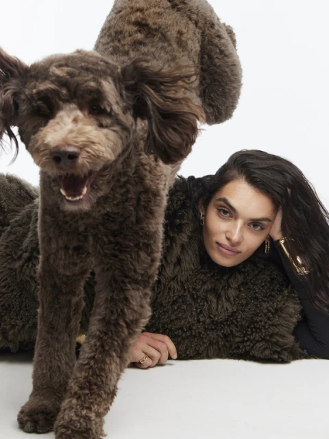 A brown poodle leaps over a light-skinned woman with long, dark hair in a studio setting, creating a dynamic and engaging portrait.