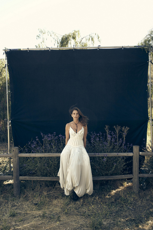 Woman in a white pleated dress sitting on a wooden fence outdoors in front of a dark screen by Victor Demarchelier