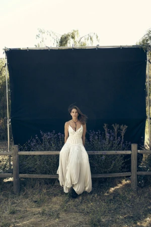 Woman in a white pleated dress sitting on a wooden fence outdoors in front of a dark screen by Victor Demarchelier