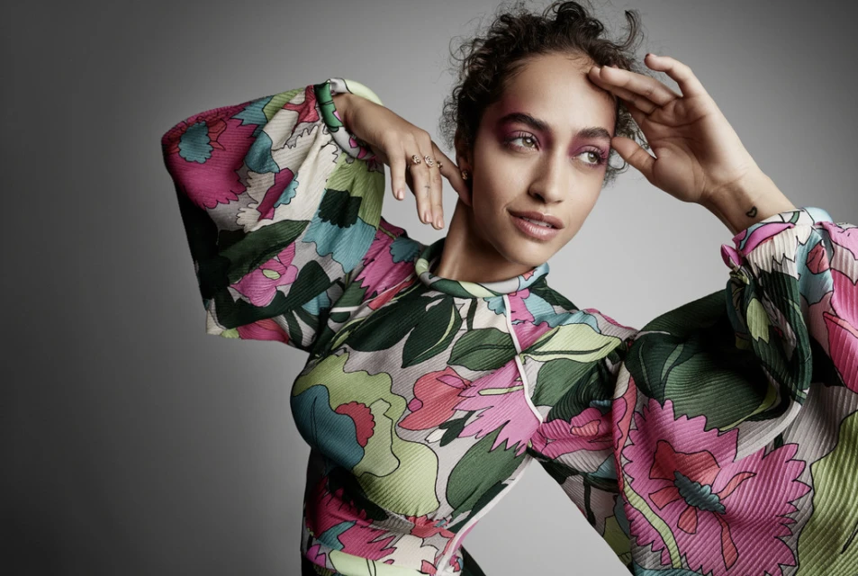 A woman poses in a floral-patterned, long-sleeved top against a grey backdrop, showcasing makeup, clothing, and studio portrait photography.