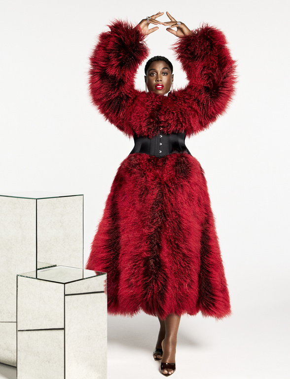 Glamorous Black woman in a red fur coat poses in a photo studio with mirrored blocks. She wears a black corset and jewelry.