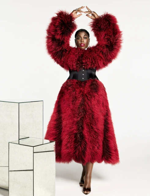 Glamorous Black woman in a red fur coat poses in a photo studio with mirrored blocks. She wears a black corset and jewelry.