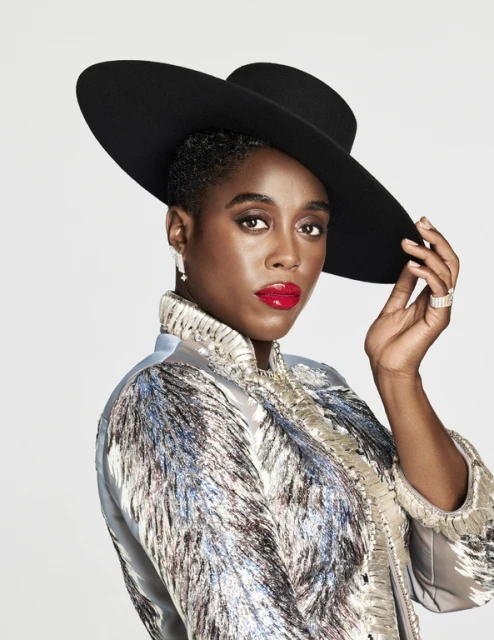 Elegant Black woman in a wide-brimmed black hat and patterned coat with red lipstick posing in front of a white background.