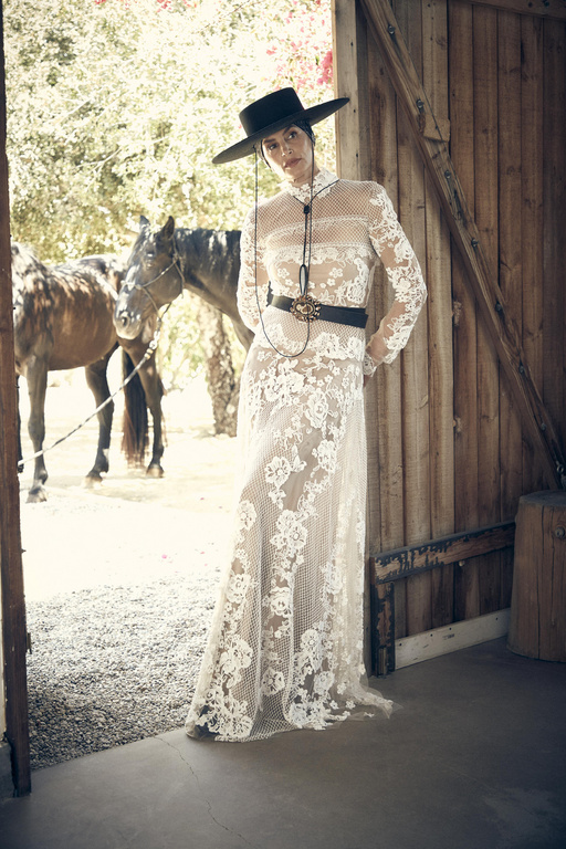 Elegant woman in a white lace dress and black cowboy hat posing in a doorway, horses in background, creating a vintage fashion vibe.