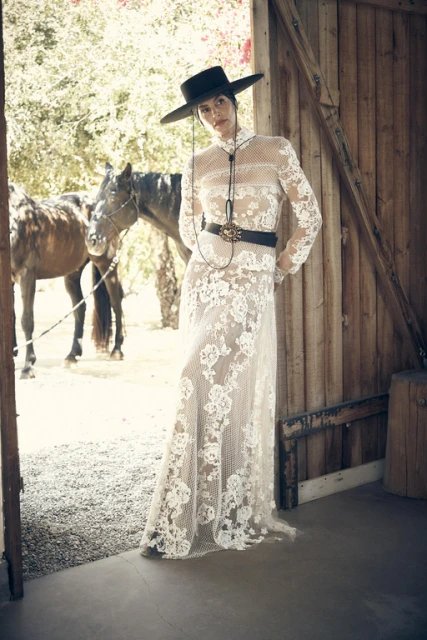 Elegant woman in a white lace dress and black cowboy hat posing in a doorway, horses in background, creating a vintage fashion vibe.