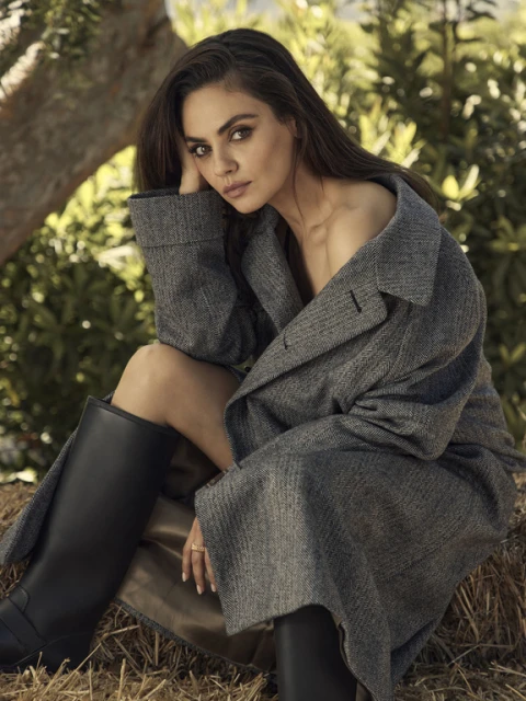 Portrait of Mila Kunis wearing a gray coat and black boots, sitting on hay outdoors with trees in the background. Fashion editorial.