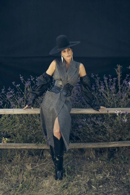 Elegant woman in a black hat and grey dress poses by a rustic wooden fence surrounded by wildflowers against a dark blue background.