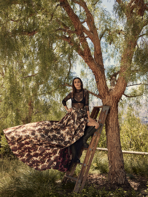 Woman in a long patterned skirt and black lace top seated on a wooden ladder beside a large tree.