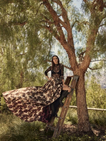Woman in a long patterned skirt and black lace top seated on a wooden ladder beside a large tree.