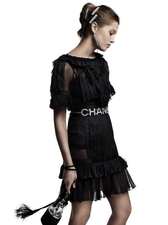 Woman in a black Chanel dress and jewelry poses against a white background. She holds a clear bottle with a black fabric covering.