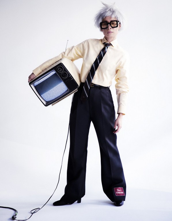 Person in retro outfit holds vintage TV with static screen in a white studio. Stylish square glasses, wig, yellow dress shirt, black tie, black pants.