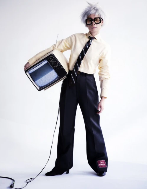 Person in retro outfit holds vintage TV with static screen in a white studio. Stylish square glasses, wig, yellow dress shirt, black tie, black pants.