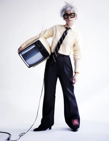 Person in retro outfit holds vintage TV with static screen in a white studio. Stylish square glasses, wig, yellow dress shirt, black tie, black pants.