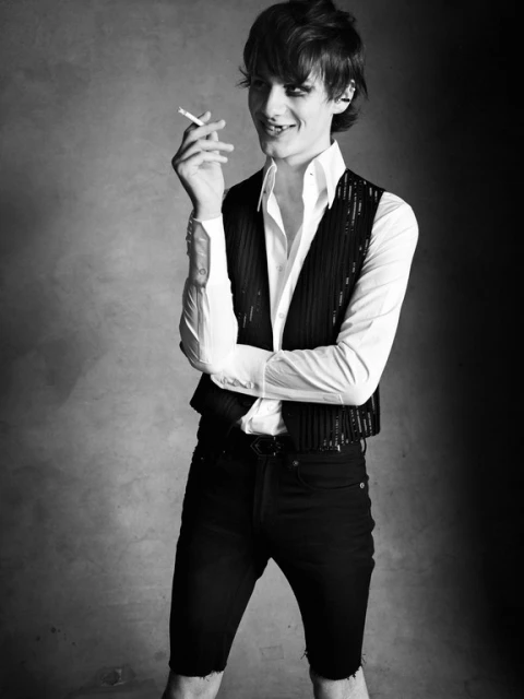 Black and white portrait of a slim adult male with dark hair, wearing a vest and shorts, and holding a cigarette, against a blurred background.