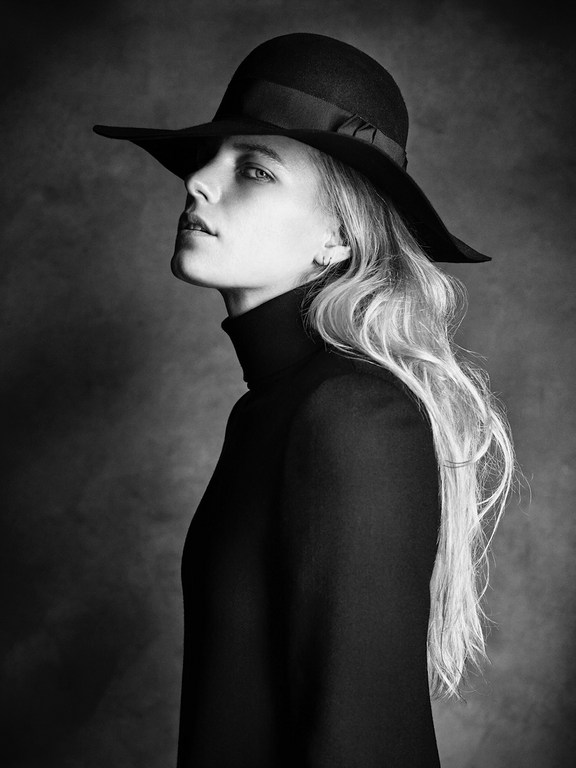 Monochrome portrait of a woman with blonde hair wearing a wide-brimmed black hat and turtleneck sweater against a dark, smoky background.
