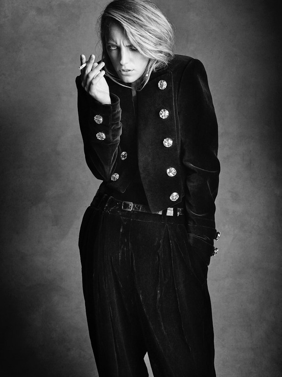 Black and white portrait of a woman in velvet jacket and pants, smoking a cigarette against a dark backdrop in a studio setting.