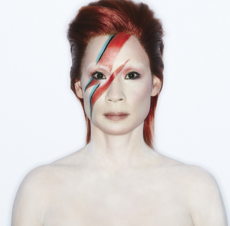 Headshot of adult woman with vibrant red hair and David Bowie-inspired face paint against a bright white backdrop in a studio setting.