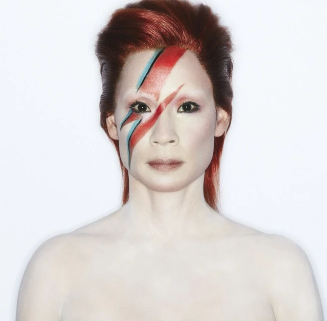 Headshot of adult woman with vibrant red hair and David Bowie-inspired face paint against a bright white backdrop in a studio setting.