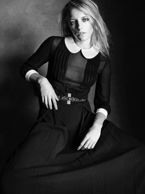 Monochrome portrait of a stylish woman with long blonde hair, wearing a sheer black blouse with a white collar and black pants.