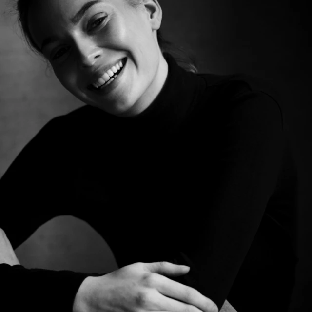 Close-up portrait of a smiling adult woman in a black turtleneck against a neutral background, shot in black and white with natural lighting.