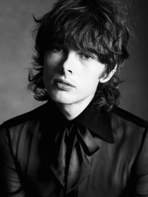 Black-and-white portrait of a stylish adult male with dark wavy hair, wearing a black blouse with a bow, in a studio setting.