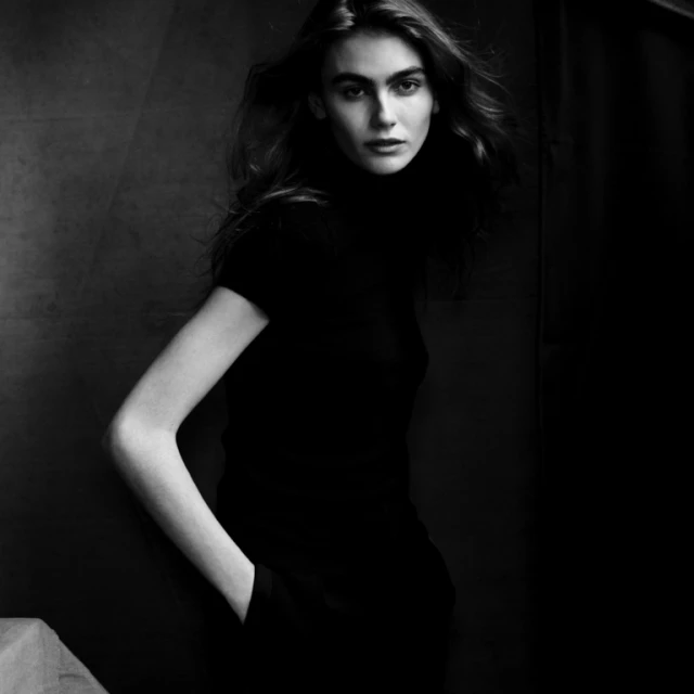 Monochrome studio portrait of a woman with long hair, wearing a black top and posing with her hand in her pocket against a dark background.