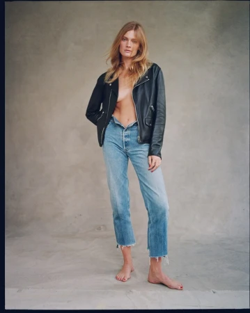 Model wearing a black leather jacket and blue jeans standing against a textured grey studio backdrop.