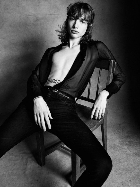 Black and white portrait of a young adult man with long hair, seated on a wooden chair and wearing an open sheer shirt and dark pants.
