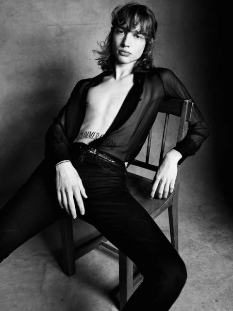 Black and white portrait of a young adult man with long hair, seated on a wooden chair and wearing an open sheer shirt and dark pants.