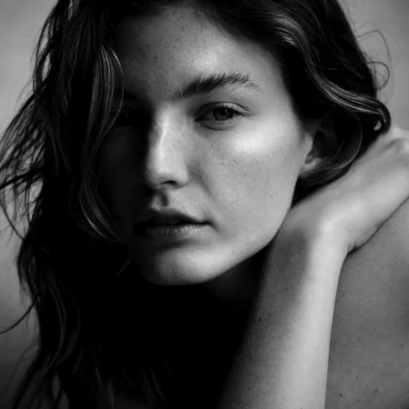 Close-up black and white portrait of a young woman with dark hair and freckles, resting her hand near her shoulder.