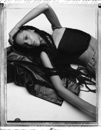 Model with long braided hair wearing a black top and leather jacket reclining on a white background