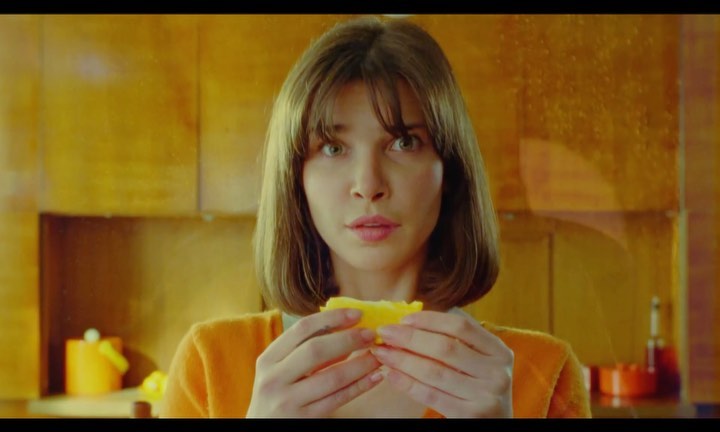Young woman in yellow sweater holds and eats a slice of lemon indoors with an expression of surprise or slight discomfort on her face.