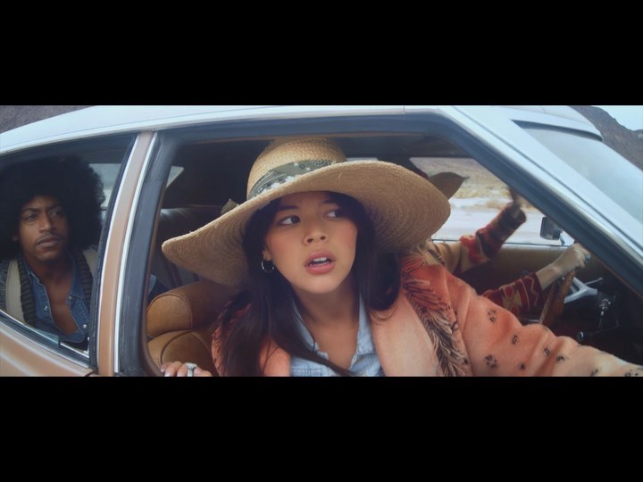 Two people inside a vintage car with desert scenery in the background. The woman wears a straw hat and patterned coat.