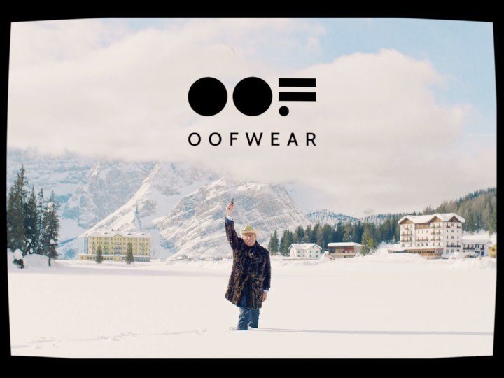 Person in fur coat raises arm in snowy field with lake and buildings in background, logo above figure, in daytime setting.