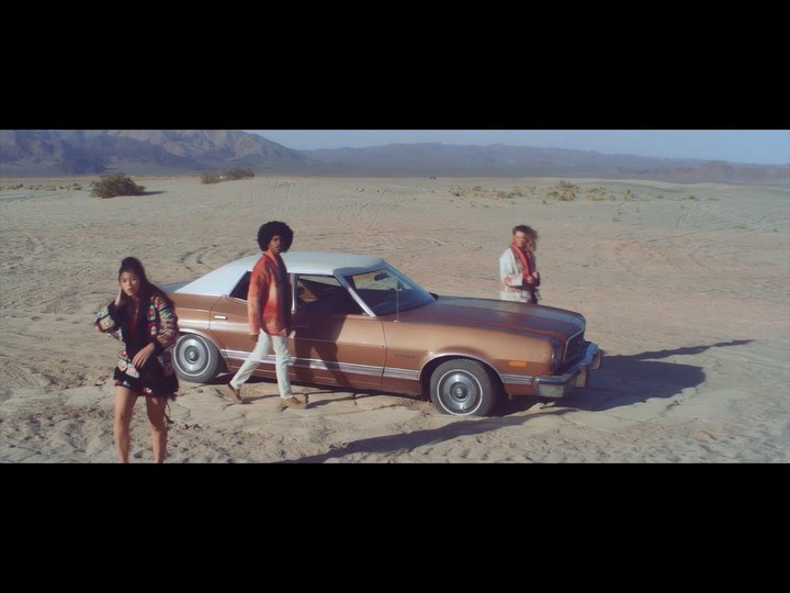 Three people stand near a vintage car in a desert landscape with mountains in the background, bathed in bright sunlight.