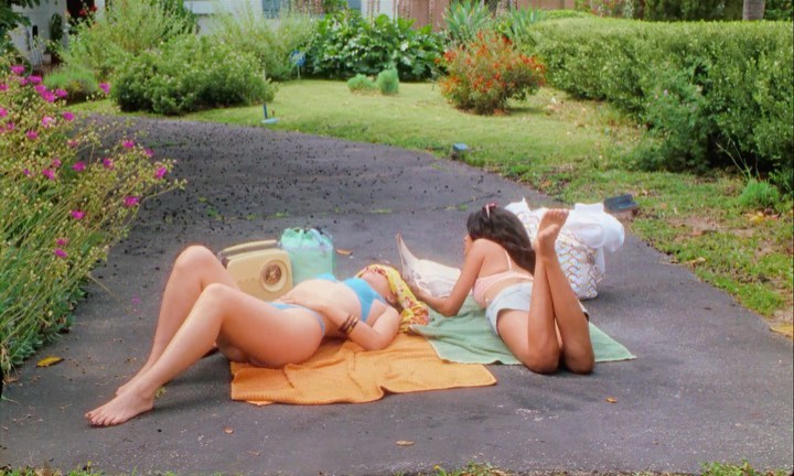 Two women in bikinis sunbathing on a driveway with a radio and bag in the background, surrounded by greenery and natural light.