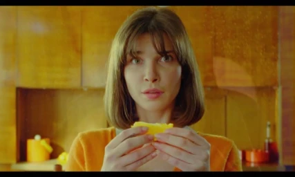 Woman with shoulder length brown hair holding a slice of orange in a domestic kitchen by Andrea Calvetti
