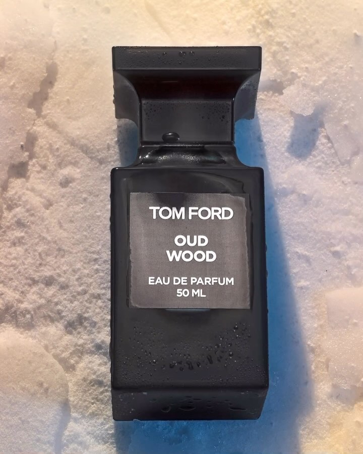 Close-up studio shot of a dark Tom Ford Oud Wood perfume bottle with a label on a textured white surface under soft, diffused lighting.