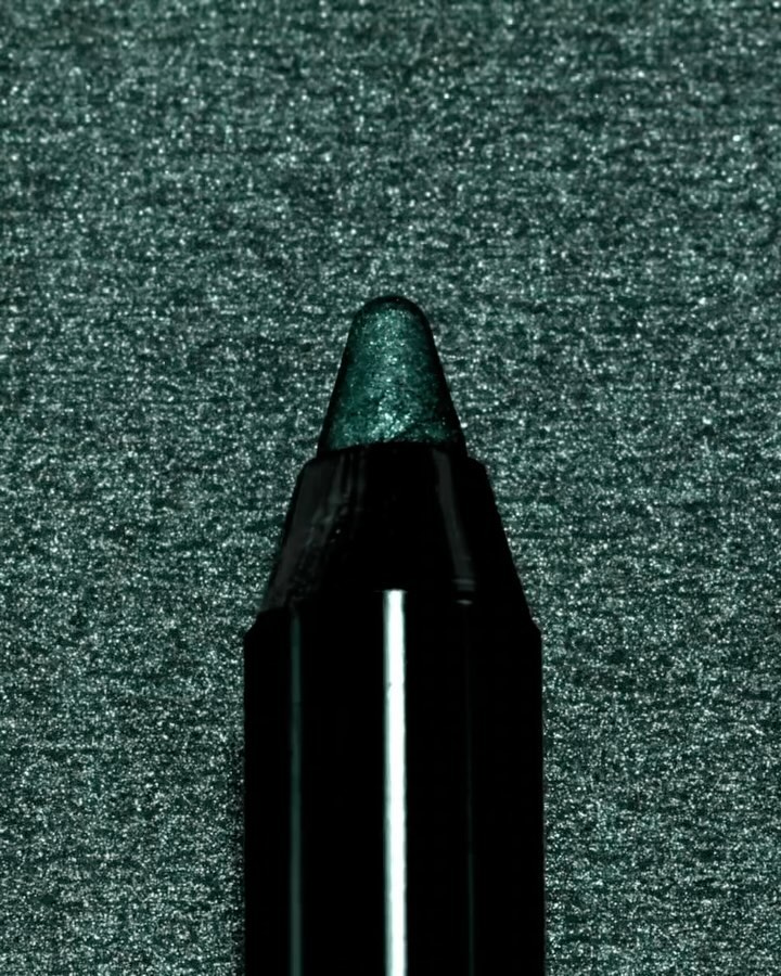 Close-up of a dark green eyeliner pencil with a shiny tip, set against a dark glittering green background.
